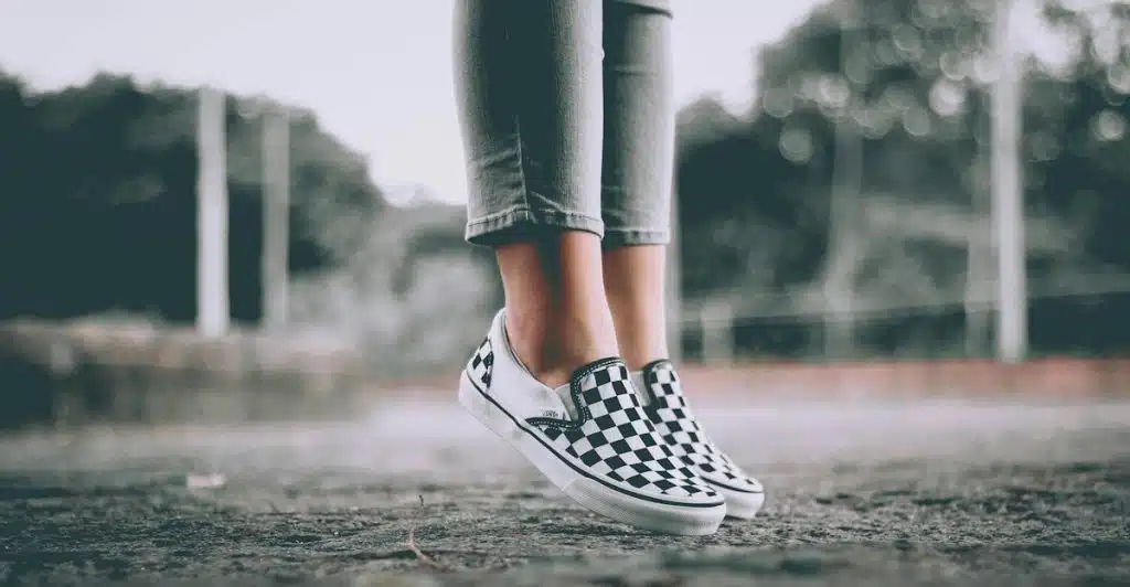 A person wearing checkered slip-on shoes and light-colored jeans standing on their toes on a path.
