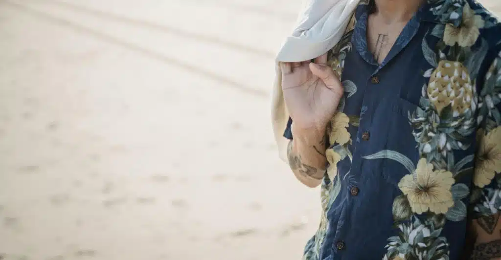 A man with tattoos wearing a dark blue floral and pineapple printed shirt with a white jacket draped over his shoulder.