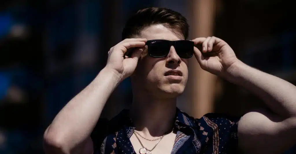 A man wearing sunglasses and a dark printed shirt with layered necklaces, adjusting his sunglasses.