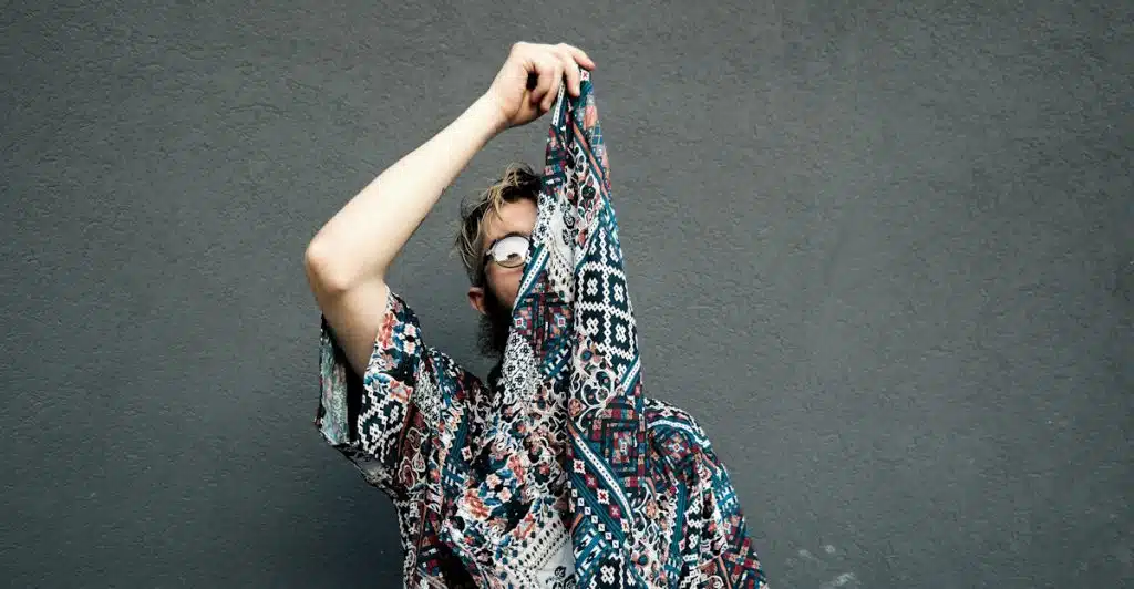 A person holding up a patterned shirt to obscure their face, standing against a dark wall.