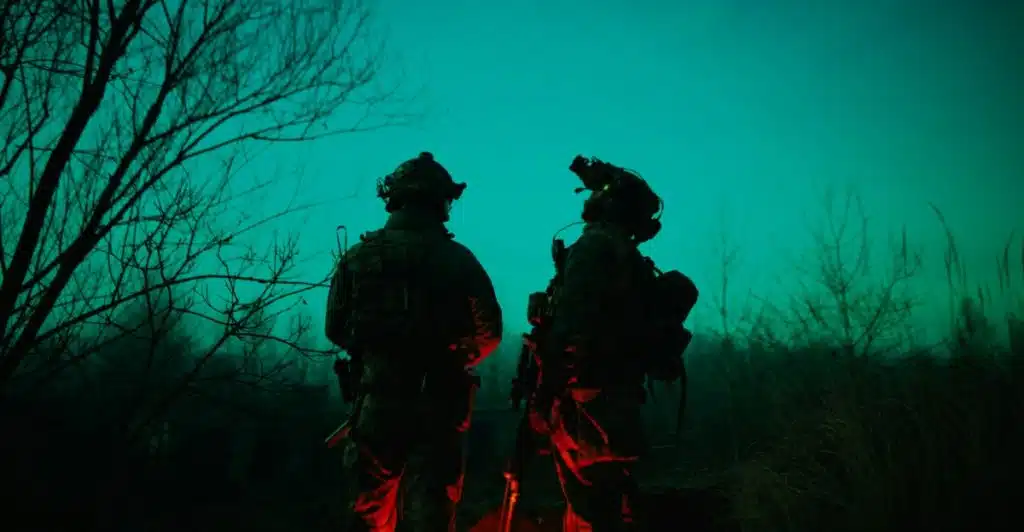 Two soldiers in tactical gear with night vision goggles standing in a dark, outdoor environment.