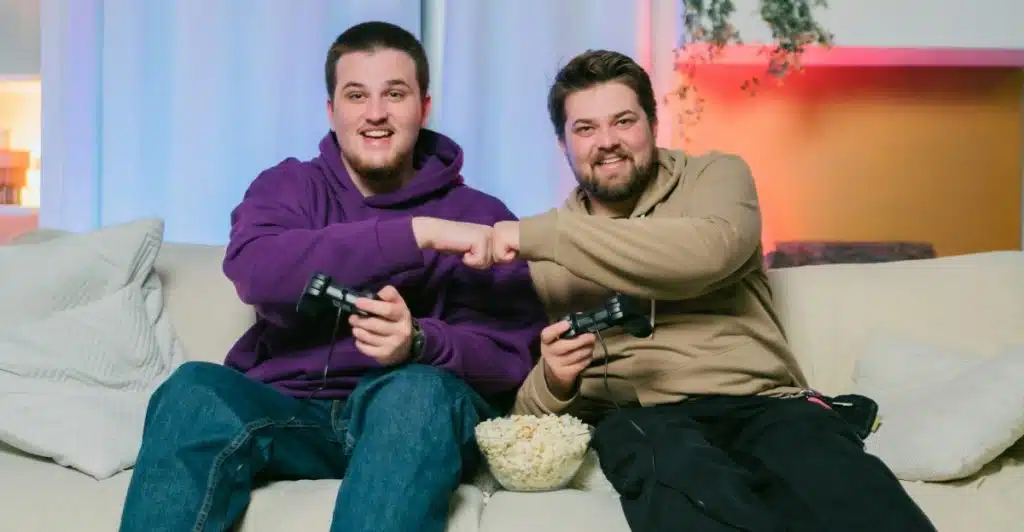 Two men sitting on a couch, one in a purple hoodie and the other in a brown hoodie, playing video games and fist-bumping with a bowl of popcorn between them.