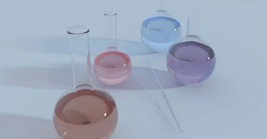 Four clear, round-bottomed laboratory flasks containing liquids of different colors along with two glass stirring rods on a light blue surface.