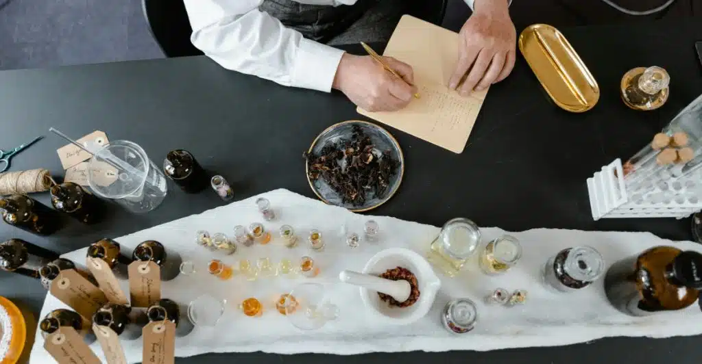 A person's hands writing in a notebook on a dark desk, surrounded by various small bottles, droppers, and dried plant materials used in fragrance creation.
