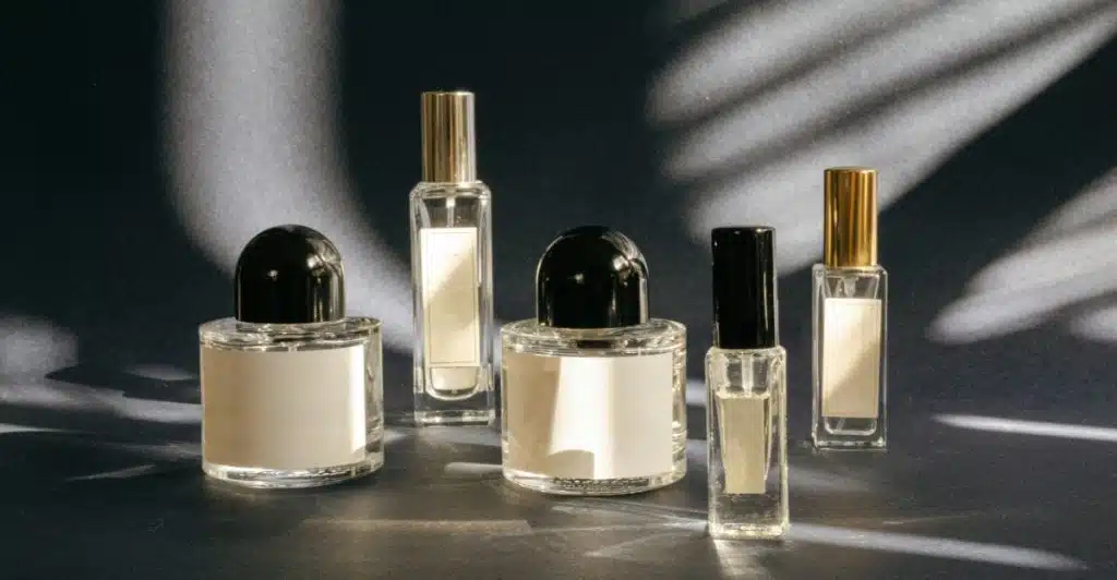 Five minimalist fragrance bottles of varying heights and shapes, with some having dark caps and some with gold, arranged on a dark, textured surface with shadows.