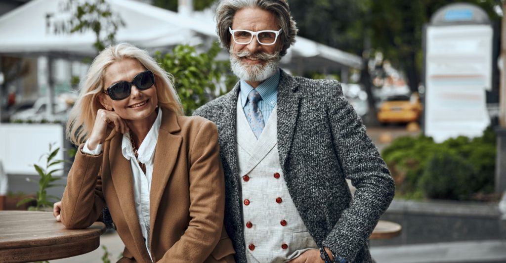 An older man with a beard and white glasses stands behind an older woman with sunglasses and a brown coat, both smiling outdoors with trees and buildings in the background.