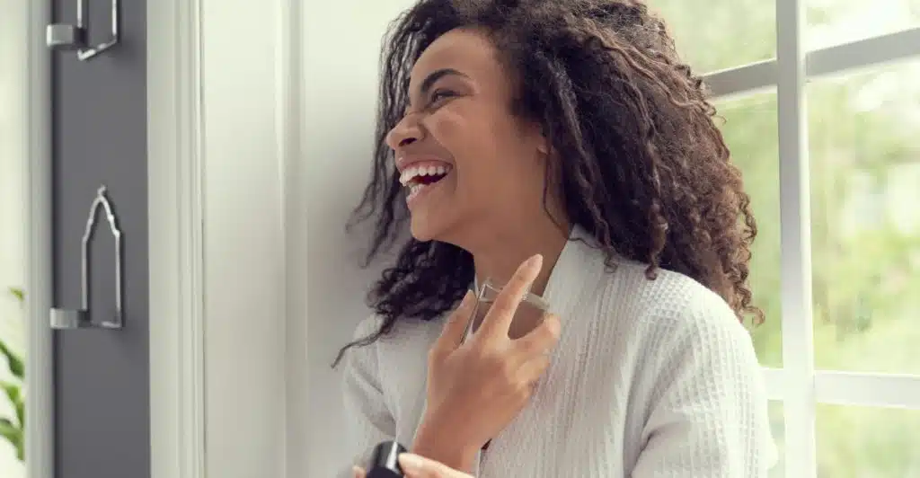 A woman with curly hair, wearing a white robe, smiles as she sprays fragrance on her neck with her eyes closed, standing near a window.