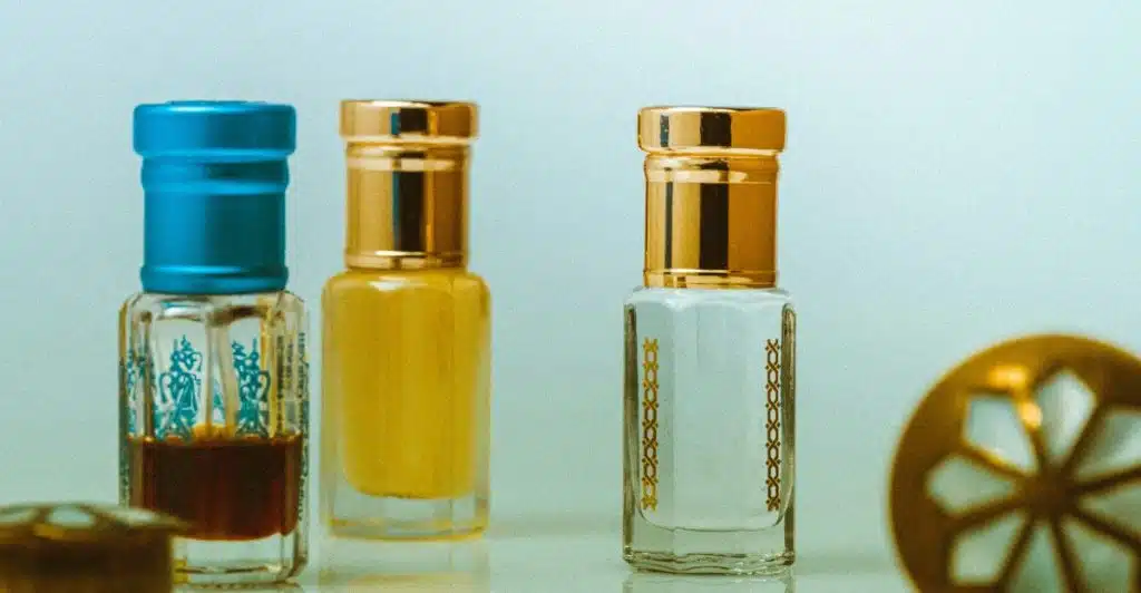  Three small, ornamental perfume sample bottles with gold and blue caps, one with dark liquid and two with lighter liquids, arranged on a light surface next to a decorative gold object.