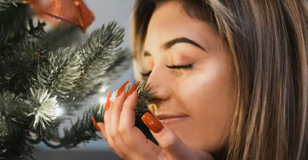 A woman with long brown hair and orange nails gently closes her eyes as she leans in to smell the branches of a decorated Christmas tree with lights.