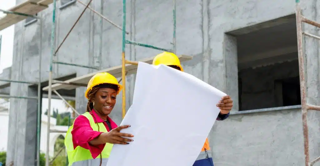 Two construction workers in hard hats reviewing blueprints at a construction site.