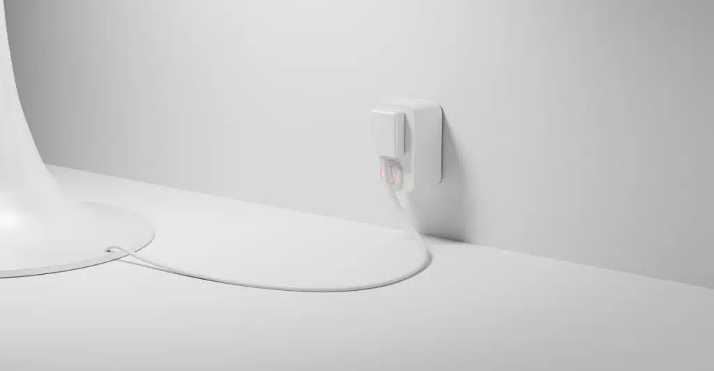 A white smart plug inserted into an outlet on a white wall, with a white cord plugged into it.