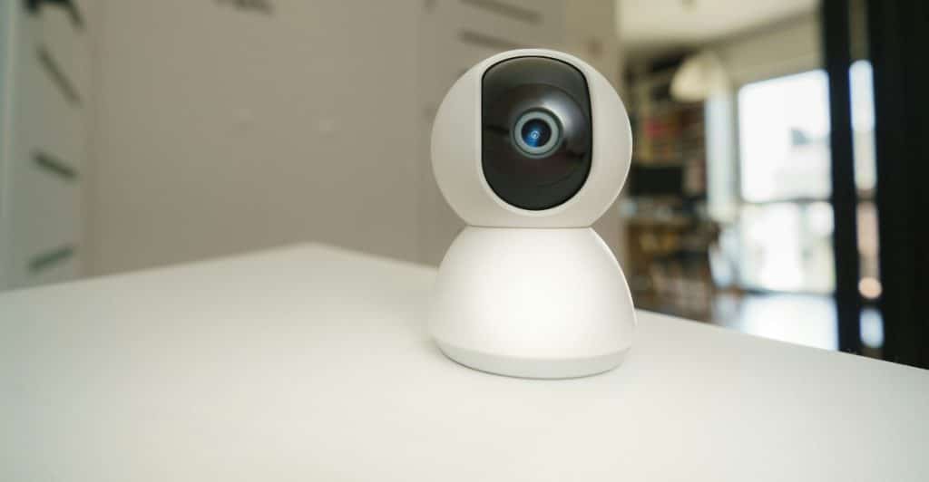 A white indoor security camera with a black lens placed on a white surface.