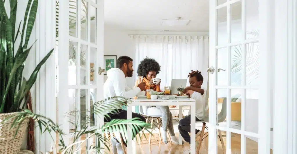 A family of three sitting at a dining table in a brightly lit room, with plants in the foreground.