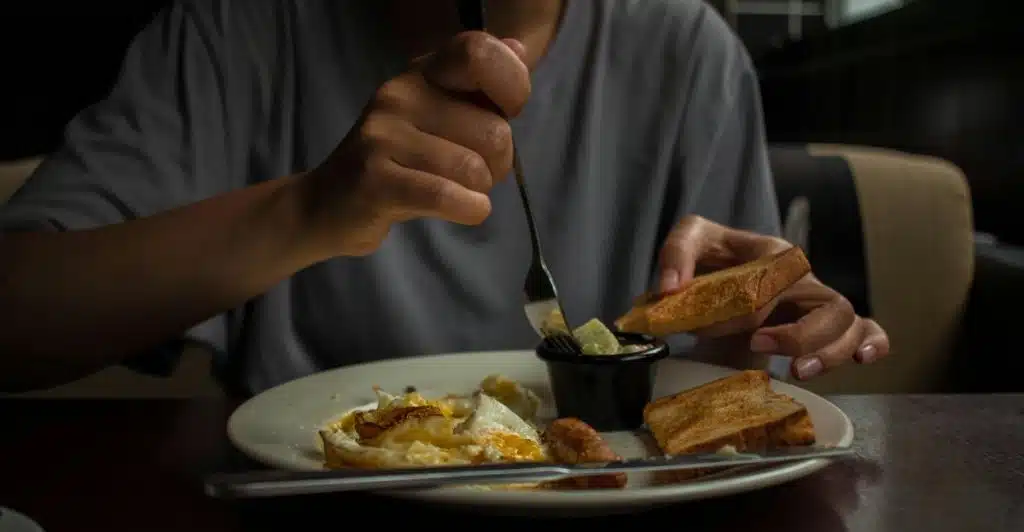 A person eating a meal with eggs and toast from a white plate at a dark wooden table.