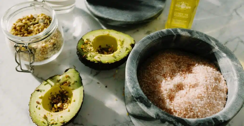 Two halves of an avocado, a jar of seeds, a mortar and pestle with salt, and a bottle of olive oil on a marble surface.
