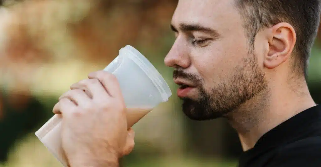 A man drinking from a white protein shaker bottle outdoors.