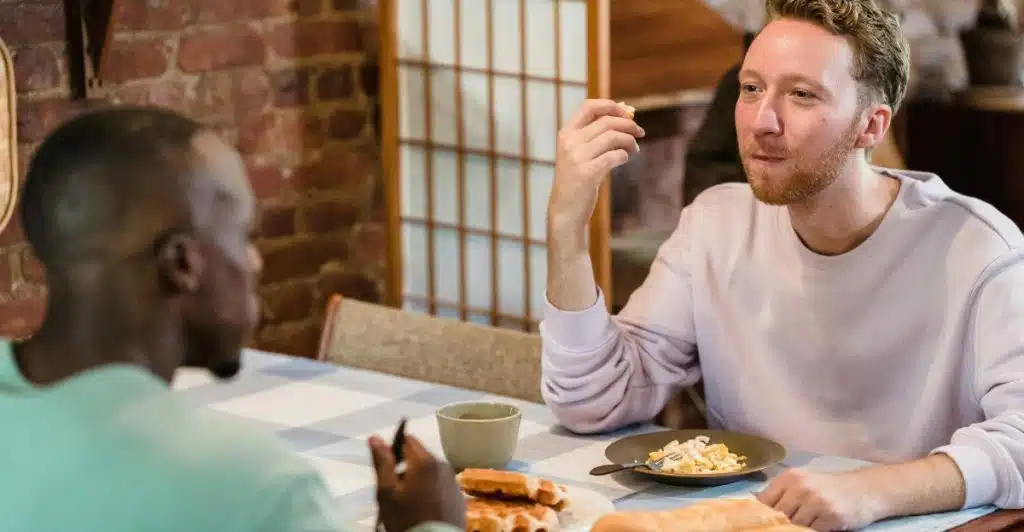 Two men sitting at a table, one eating and the other looking at a phone, with plates of food and cups.