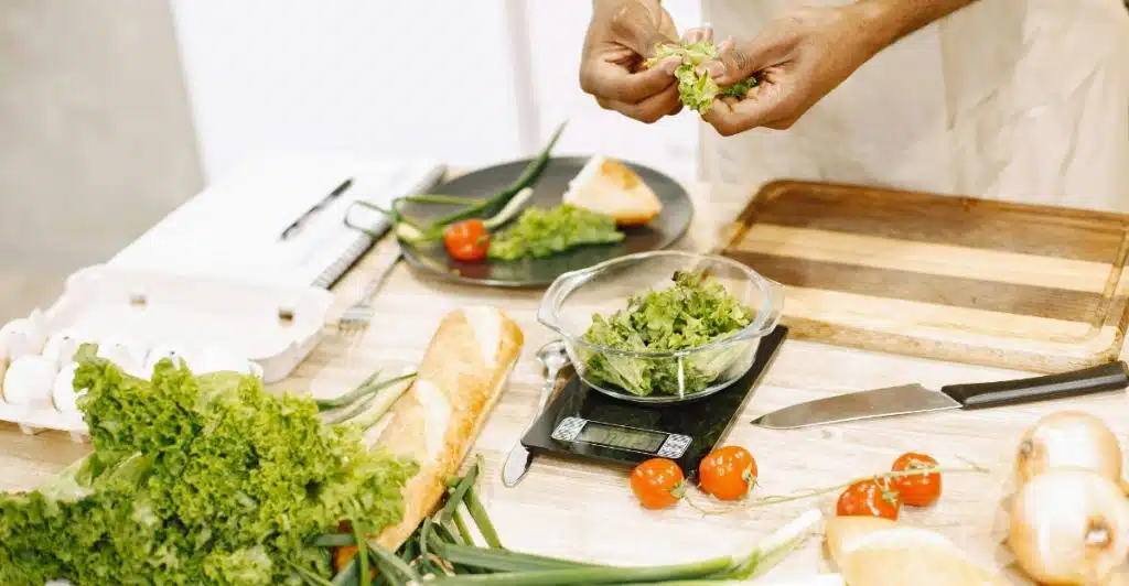 A man in an apron preparing vegetables and other ingredients on a wooden counter with a food scale.