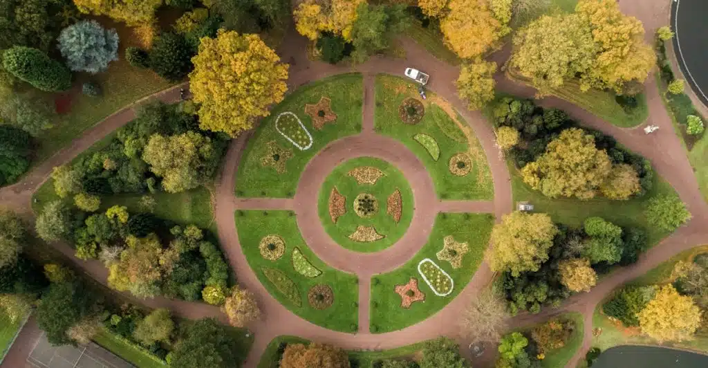 An aerial view shows a circular park design with pathways, various green and yellow trees, and sections of manicured gardens, with a small body of water and tennis courts visible at the edges.