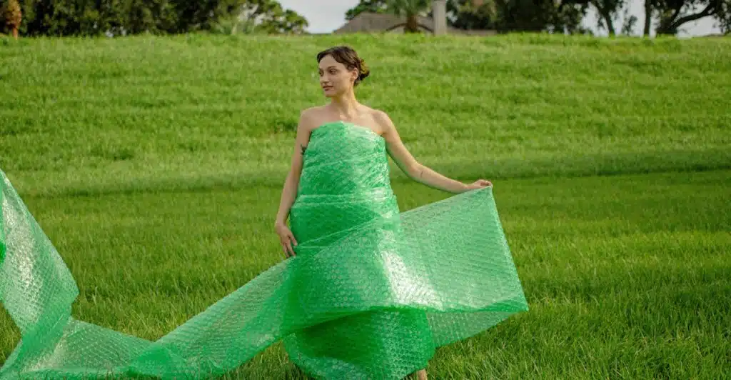 A woman with dark hair wearing a long, flowing green dress made of bubble wrap stands in a lush green grassy field, looking off to the side.