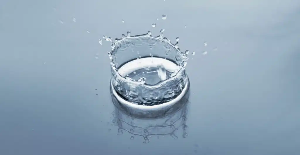 A single water droplet creates a crown-like splash as it lands on a smooth, reflective light blue surface.