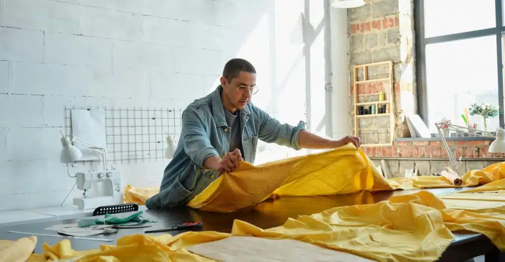 A man wearing glasses and a denim jacket is carefully folding a large piece of bright yellow fabric on a wooden table in a well-lit room with sewing machines in the background.