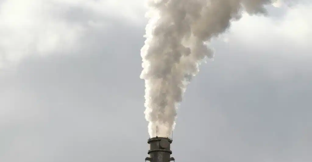 A tall, dark industrial smokestack is emitting a large, thick plume of white smoke or steam into a hazy sky.