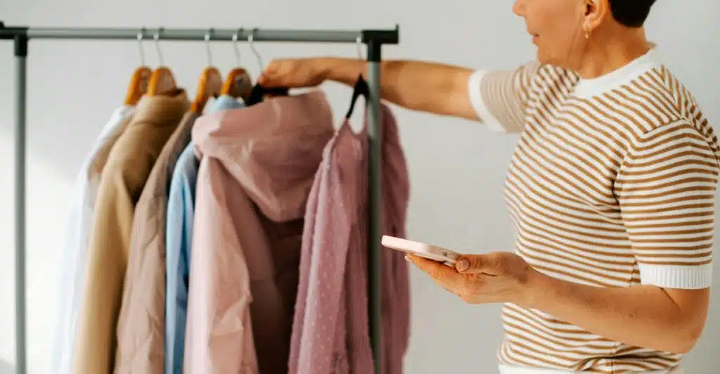 A person is standing in front of a clothing rack filled with various garments, holding a smartphone in their hand, with more clothes visible on another rack in the background.