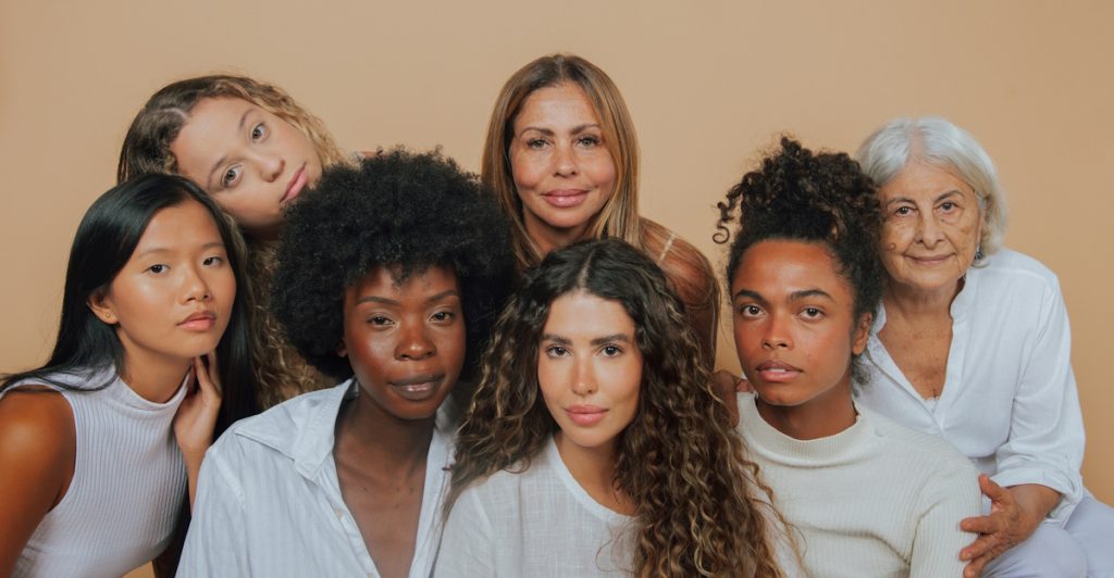 A group of six women with diverse ages and appearances are posing together, some smiling and some looking towards the viewer, against a plain light brown background.