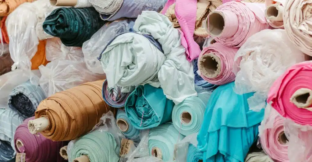 Various rolls of colorful fabric stacked together, including shades of pink, teal, mint, and orange, wrapped in plastic and stored in a textile market or warehouse.