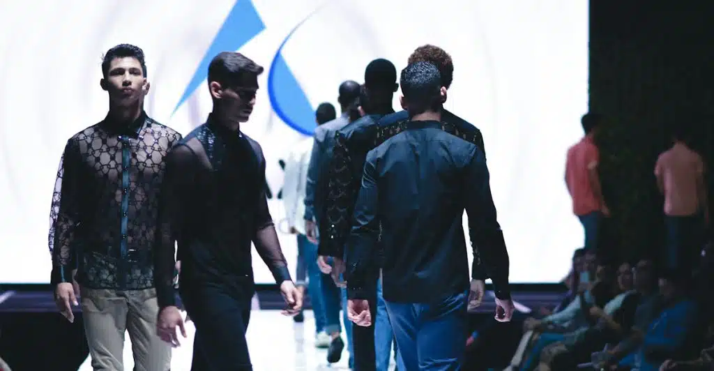 Several male models are walking down a brightly lit fashion runway, showcasing various dark-colored outfits, with an audience seated on the sides.
