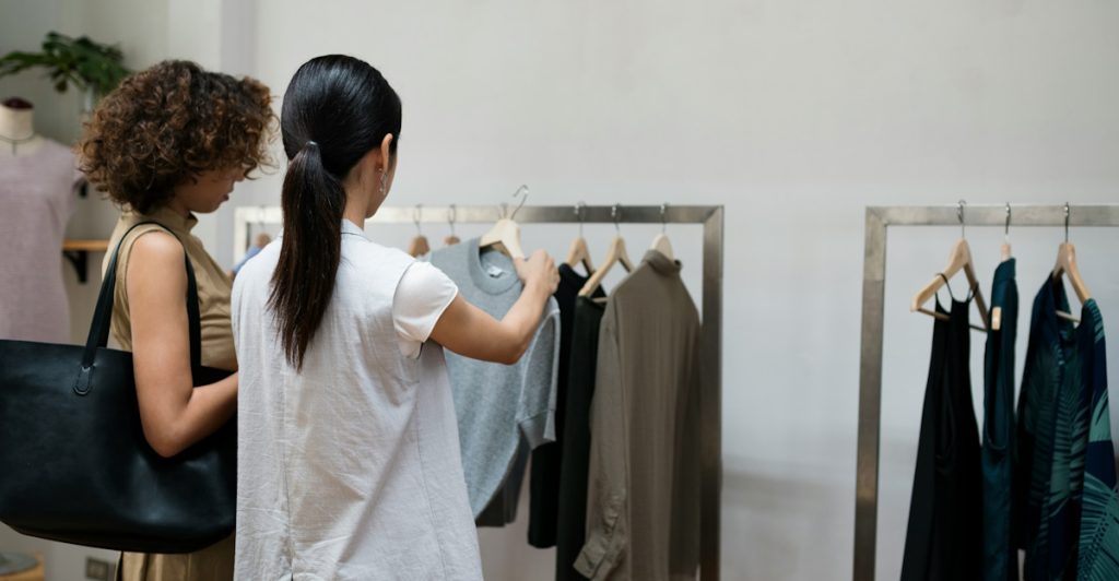 Two women are standing in a clothing store, with one woman facing a rack of clothes and the other woman looking on, surrounded by various garments on hangers.