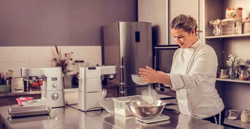 A woman in a chef's coat sifting powder into a bowl in a professional kitchen.