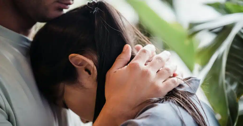 A man embracing a woman, holding her head to his chest with his hands on her shoulders and neck.