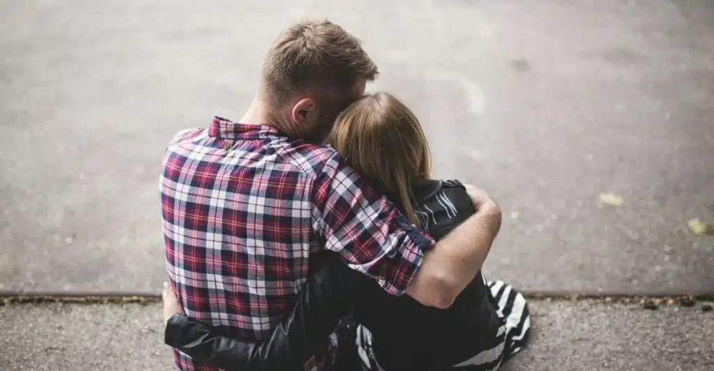 A man and a woman are sitting on the ground with their backs to the camera, embracing each other.