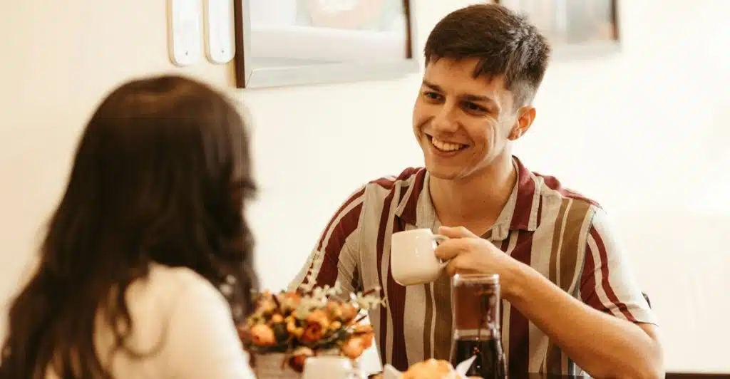 A man and a woman are seated at a table in a cafe, looking at each other and smiling while the man holds a coffee cup.