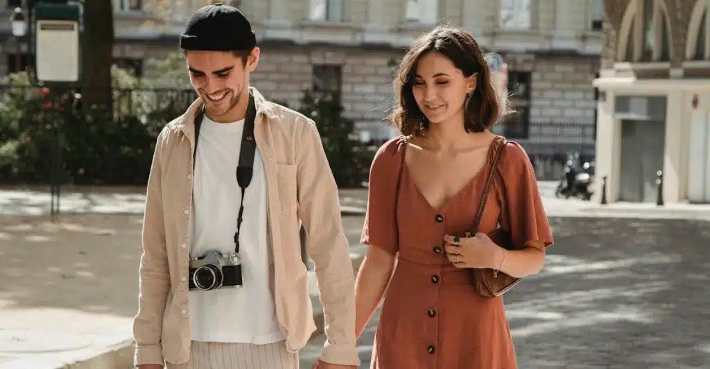 A man and a woman walking outdoors, holding hands and looking down.