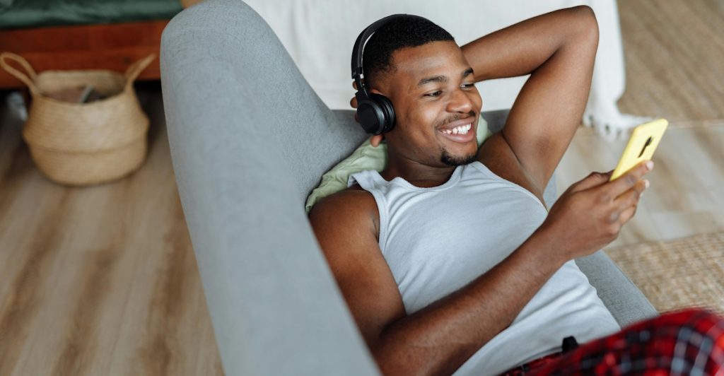 A man is reclining on a chair, wearing headphones and looking at his phone.