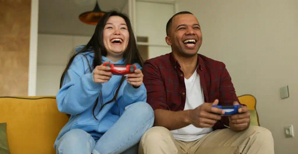 A man and a woman sitting on a couch, laughing while holding video game controllers.