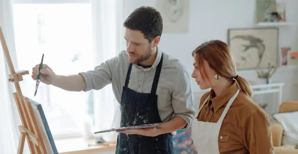A man and a woman are standing in front of an easel, with the man holding a paintbrush and looking at the canvas.