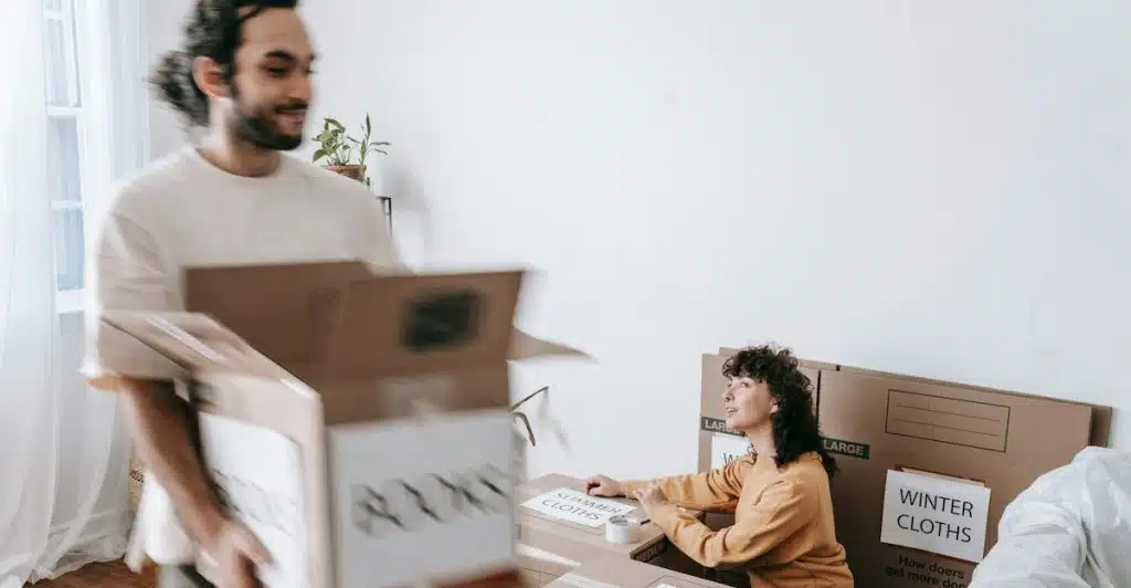 A man carrying a box while a woman sits surrounded by other boxes, appearing to be moving.