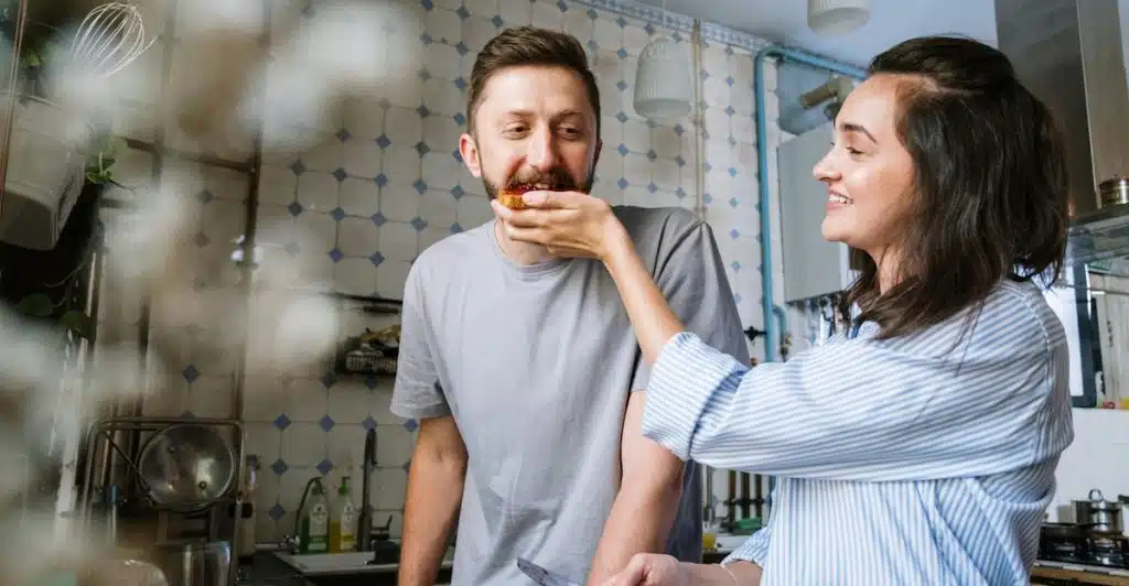 A woman feeding a piece of food to a man in a kitchen.