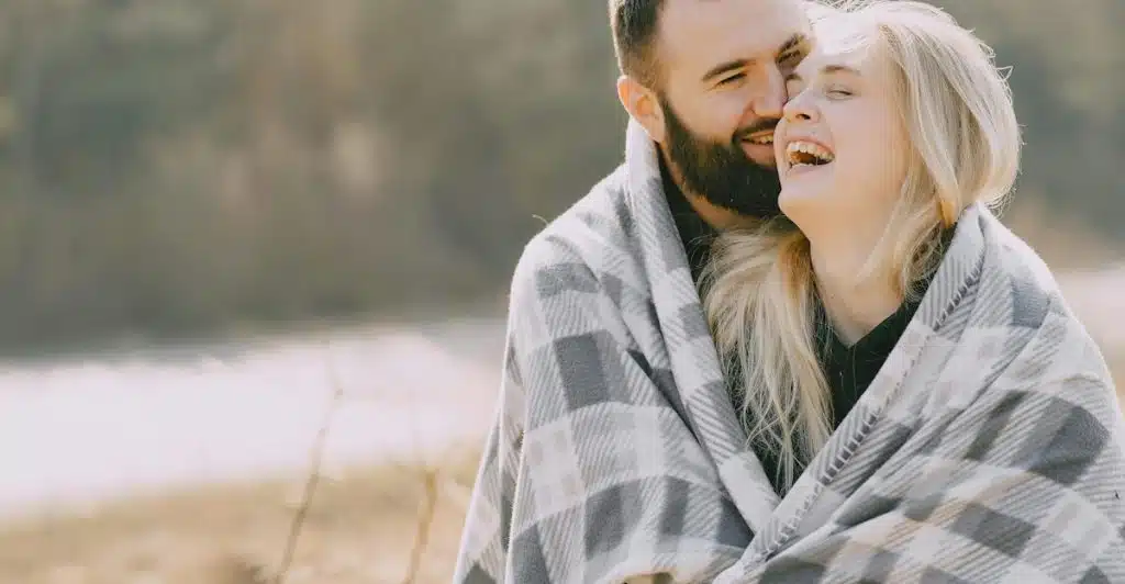 A man and a woman wrapped together in a plaid blanket outdoors, smiling and laughing.