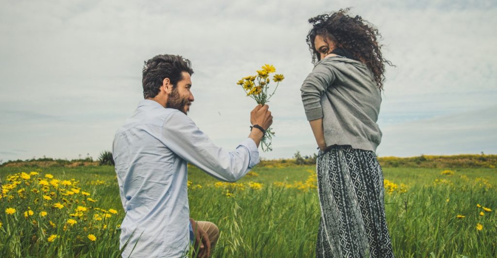 A man kneeling in a field of yellow flowers, offering a bouquet to a woman standing with her back partially turned,