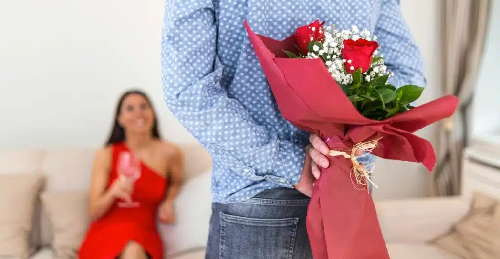 A man holding a bouquet of red roses behind him, with a woman in a red dress smiling in the background