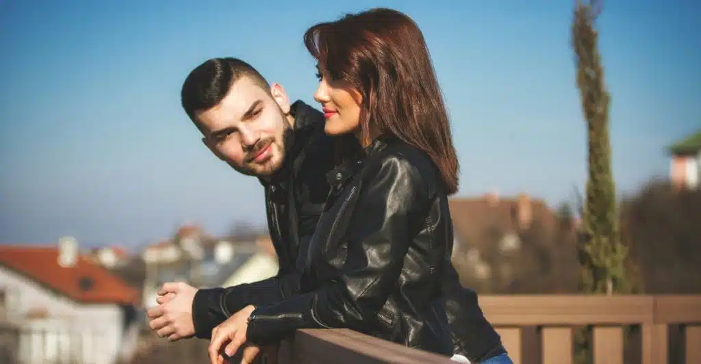 A man and a woman in leather jackets leaning on a wooden railing, with the man looking at the woman and smiling.
