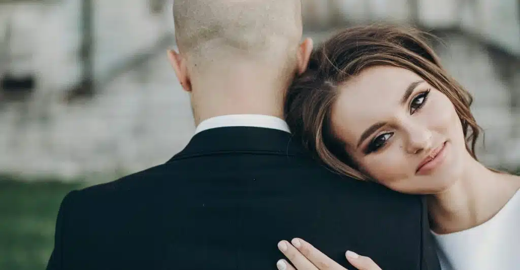 A woman in a white dress with her head resting on a man's shoulder, seen from behind him.