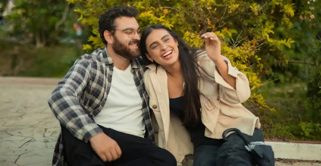 A man with glasses and a plaid shirt sitting next to a woman with long dark hair, both laughing outdoors.