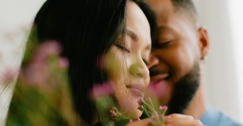 A man embracing a woman from behind, both with their eyes closed, with purple flowers in the foreground