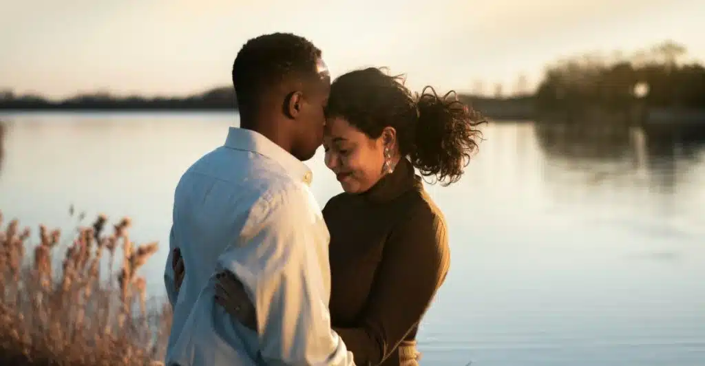 A man and a woman embracing by a lake, with the man kissing the woman's forehead.